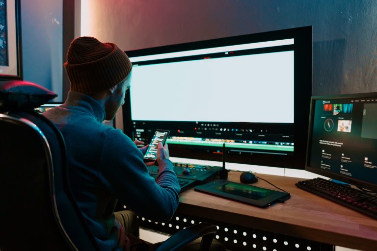 Male Video Editor Works with Footage or Video on His Personal Computer and having a break communicating on his smartphone. He Works in Creative Office Studio or home.